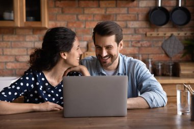 Modern mutfakta dizüstü bilgisayar kullanan mutlu genç çift, ahşap masada oturuyor, aletlerle evde eğleniyor, gülümseyen adam ve kadın online alışveriş yapıyor, internette sörf yapıyor, komik videolar seyrediyor.