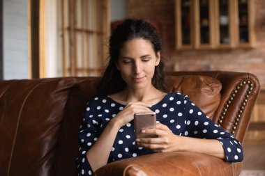 Akıllı telefon kullanan, uygulamaları tarayan, evdeki rahat koltukta tek başına oturan, hoş, genç bir bayan telefon ekranına bakıyor, sosyal ağlarda sohbet ediyor, online alışveriş yapıyor, video izliyor.