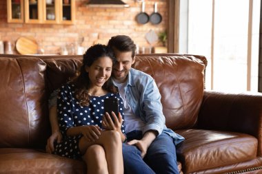 Mutlu genç çift sarılıyor, akıllı telefonunla birlikte eğleniyor rahat rahat koltukta oturuyor, gülümsüyor heyecanlı kadın ve erkek telefon ekranına bakıyor video görüşmesi yapıyor, sohbet ediyor ya da internetten alışveriş yapıyor.