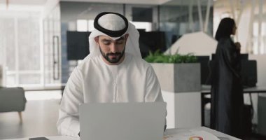 Genç 35 'lik Arap işadamı geleneksel Müslüman beyaz giysileri içinde çağdaş ofis bilgisayarında çalışıyor, müşteri e-postasına cevap veriyor, gelen uyarıyı okuyor, gülümsüyor, cevap veriyor, güncellemeleri gözden geçiriyor