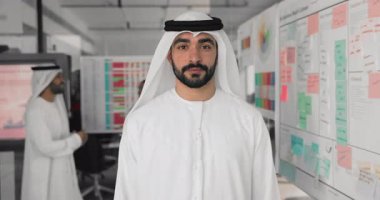 Beyaz kandora gibi giyinmiş Arap işadamı ya da yönetici, kollarını kavuşturmuş duruyor, ofiste kendinden emin bir gülümsemeyle kameraya bakıyor, stratejik iş yönetimini temsil ediyor, liderliği yayıyor.