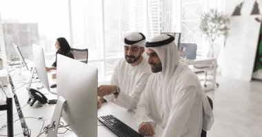 Yeni iş platformunu inceleyen iki Arap erkek meslektaşı, yatırım olanakları veya çağdaş çalışma ofisindeki bilgisayarla masada oturan proje güncellemeleri. İşbirliği, modern teknoloji