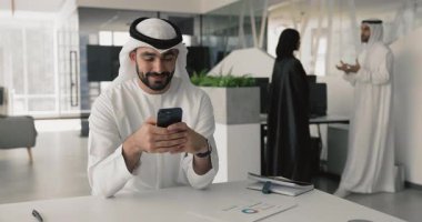 Beyaz kandora giymiş, akıllı telefon kullanan Arap bir adam modern iş yerinde masa başında oturuyor, müşteriyle dijital profesyonel iletişim kuruyor, mesajlarını paylaşıyor, tatilde kişisel sohbetlerini yönetiyor.