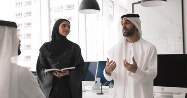 Geleneksel beyaz kandura ve ghutra 'lı bir adam, modern çalışma ofisinde duran meslektaşlarıyla brifing sırasında fikir sunuyor ya da açıklama yapıyor. İşbirliği, proje planlaması