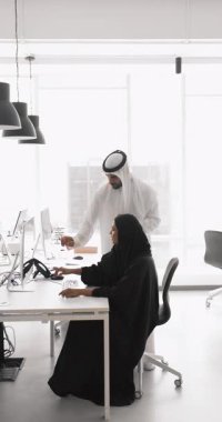 Beyaz kandoralı genç Arap bir adam, masa başında oturmuş, yeni program ya da yazılıma bakarak görev ya da iş platformuna yardım ediyor. Akıl hocalığı, çıraklık, takım çalışması.