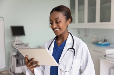 Neşeli genç siyahi doktor kadın tablet üzerinde elektronik tıp raporu okuyor, internet iletişim teknolojisi kullanıyor, tıbbi başvuru yapıyor, kayıtları kontrol ediyor.