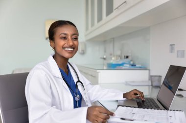Neşeli genç Afrikalı Amerikalı pratisyen, dizüstü bilgisayarda yazıyor, hastane ofisindeki tıbbi kağıt kayıtlarını inceliyor, başka tarafa bakıyor, gülümsüyor, gülüyor. Mutlu doktor kadın samimi bir portre