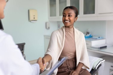 Sağlıklı, genç Afrikalı bir hastanın, kadın doktora minnettarlıkla el sıkışması, mutlu bir gülümseme, klinik muayene koltuğunda oturması, tıbbi profesyonele teşekkür etmesi, el sıkışması.