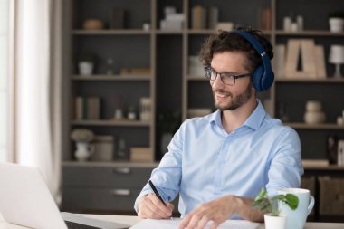 Motive edilmiş uzaktan kumandalı öğrenci. Kablosuz stereo kulaklık takan yeniyetme meraklı genç adam not defteriyle e-öğrenme platformu ile ilgili interaktif çevrimiçi ders derslerine katılıyor.