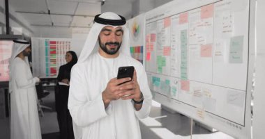 Genç Arap iş adamı, beyaz bulaşık makinesi giymiş ofis çalışanı iş yerinde akıllı telefon kullanıyor, mesajları paylaşıyor, e-postayla cevap veriyor, gülümsüyor, uzaktan dijital iş iletişiminden tatmin oluyor.