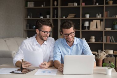 Gülümseyen iki genç kardeş tek yumurta ikizi işadamları girişimciler ofis masasında evrak işlerinde işbirliği yaparak pazarlama istatistiklerini analiz ediyorlar not defteri tableti kullanarak projeyi hazırlıyorlar..