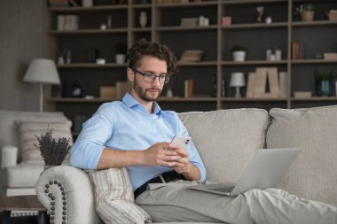 Modern çağda birden fazla iş. Rahat genç erkek kanepede dinlenir cep telefonu ve not defteri arasında dosya alışverişi sağlar hücre uygulamasında kolay veri senkronizasyonu için Platformlar arasında bilgi erişimi için metin mesajı