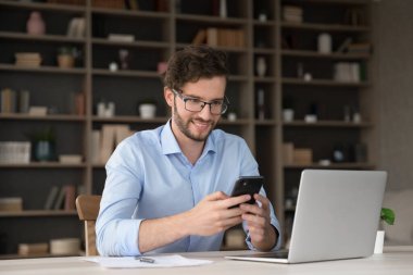 Kolay ve verimli bir çoklu görev. Gülümseyen genç adam uzaktan çalışan bir çalışan iş yerinde bilgisayar başında oturuyor telefon uygulaması ile etkileşime geçip mesai arasında sosyal medyayı kontrol etmek için iş arkadaşına mesaj gönderiyor.