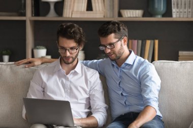 Dijital işbirliği. Dost canlısı genç ikiz kardeşler kanepede oturur online eğitim için defter kullanırlar serbest çalışma eğlence ekibi ekrana bakan beyin fırtınası fikirleri üzerinde çalışırlar.