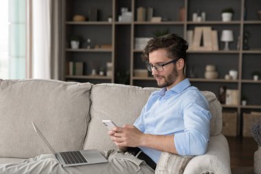 Dijital yaşam tarzı. Rahatlamış erkek milenyum zamanı evde rahat bir kanepede oturarak akıllı telefondaki cihazlar ile Wi-Fi üzerinden iki aygıt arasındaki bilgisayar paylaşımı bilgisinden dikkati dağıtılmış mesajlar kullanıyor.