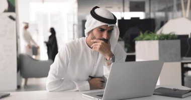 Kandura gibi giyinmiş Arap bir adam ofiste proje üzerinde çalışıyor, akıllı telefon ve kağıtla masaya oturuyor, bilgileri gözden geçiriyor, alınan verileri inceliyor, istatistikleri analiz ediyor, beyin fırtınası fikirlerini inceliyor, arama çözümünü araştırıyor.
