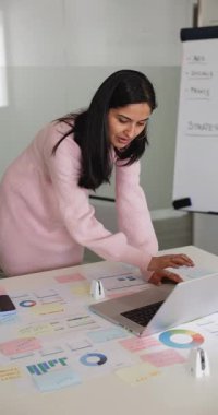 Ofiste çalışan Hintli bir kadın, dizüstü bilgisayarda yazı yazıyor. Etrafı çizelge ve şemalarla çevrili. Rapor ya da sunum hazırlıyor, verileri gözden geçiriyor, iş e-postalarına cevap veriyor.