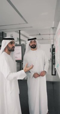 Geleneksel beyaz Müslüman elbiseli iki genç Arap meslektaşı, duvara iliştirilmiş bilgileri gözden geçiriyor, ortak proje güncellemelerini tartışıyor, fikirleri paylaşıyor, yaratıcı çözümleri veya teslim tarihlerini paylaşıyor