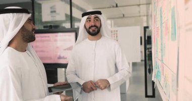 Geleneksel beyaz kandura giymiş iki Arap, modern ofiste duvarın yanında yapışkan notlarla duruyor, yol haritasını, görsel tahtaları gözden geçiriyor, takım hedeflerini analiz ediyor, plan stratejisini inceliyor.