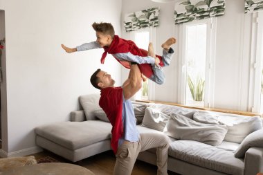 Kırmızı pelerinli güçlü baba ve oğul evde yaratıcı bir süper kahraman oyunu oynuyor. Baba oğlunu uçan bir süper kahraman gibi havaya kaldırıyor, çocuk kollarını öne doğru açıyor, uçan bir poz gibi.