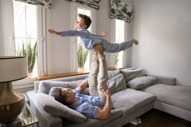 Oturma odasındaki kanepede uzanmış genç ve güçlü bir baba, uçarken küçük 8 'li yaşlarındaki oğlunu havaya kaldırıyor, uçuyormuş gibi yapıyor, kollarını uçak kanatları gibi açıyor. Aile oyunları, destek, aktif, keyifli ev eğlencesi