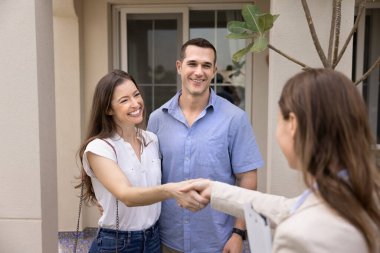 Evli genç çift, kadın emlakçıyla el sıkışıyor, modern kır evinin verandasında yan yana duruyorlar. Gayrimenkul işlemleri, ev sahipliği ve profesyonel danışmanlık