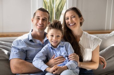 Küçük oğlu kameraya poz veren neşeli bir çiftin portresi rahat koltukta oturuyor, mutluluk saçıyor, sıcaklığı paylaşıyor, sevgiyi paylaşıyor, modern dairede birlikte paha biçilmez zaman geçiriyorlar. Kira, aile bağları