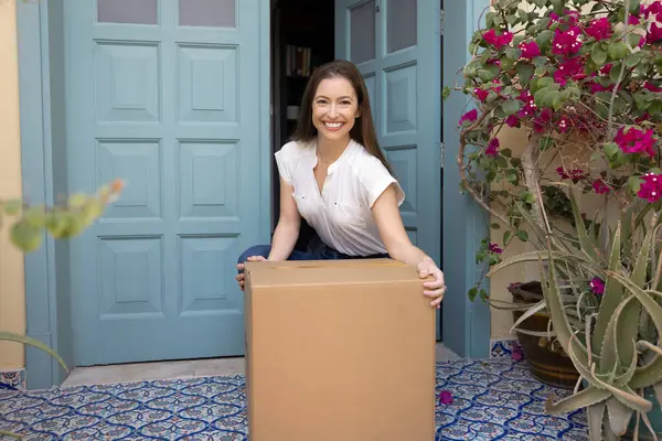 Evinin girişinde diz çökmüş gülümseyen genç bir kadının portresi, büyük bir karton paketi tutmak, başarılı teslimat hizmetlerinden tatmin olmak, paket kutusunu açma beklentisi