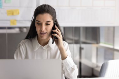 Odaklanmış genç İspanyol iş kadını iş yerinde iş iletişimi için aletler kullanıyor, cep telefonuyla ve dizüstü bilgisayarla konuşuyor, cep telefonuyla pazarlık yapıyor, cep telefonuna danışmanlık yapıyor.