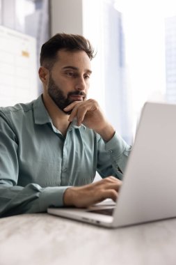 Ofiste dizüstü bilgisayarda çalışan düşünceli, yakışıklı Latin iş adamı, derin düşüncelerde yüze ve çeneye dokunuyor, online satış büyümesi, internet pazarlama stratejisi üzerinde düşünüyor. Dikey çekim