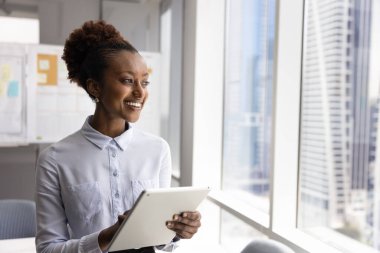 Ofiste dijital tablet kullanan mutlu, genç Afrikalı iş kadını pencereden dışarı bakıyor, gülümsüyor, başarılı bir kariyer düşünüyor, internet pazarlama stratejisi düşünüyor.