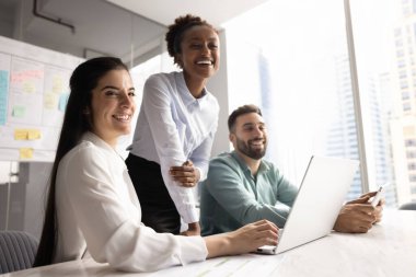 Neşeli ofis arkadaşları toplantı odasında takım çalışmasının keyfini çıkarıyorlar. Masada birlikte dizüstü bilgisayar kullanıyorlar. Başka tarafa bakıyorlar, ekranın dışında iş arkadaşlarıyla konuşuyorlar.