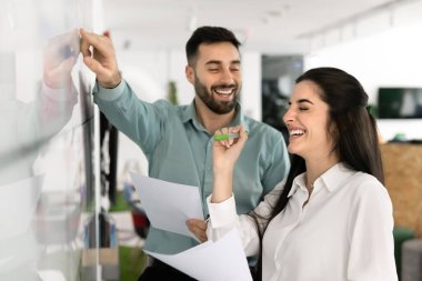 Neşeli iş projesi meslektaşları eğleniyor, beyaz tahtaya işaret ediyor ve gülüyor, takım çalışmasından keyif alıyor, görev programlama, başlangıç yönetimi üzerinde birlikte çalışıyorlar.