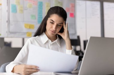 Düşünceli Latin iş kadını iş yerinde finans belgesi okuyor, kafaya dokunuyor, problem çözmeyi düşünüyor, satış pazarlama raporunu analiz ediyor, kağıt faturasını kesiyor.