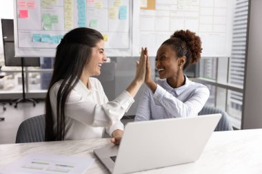 Heyecanlı çeşitli kadın iş arkadaşları ofis masasında beşlik çakıyor, başarılı bir işbirliği için dizüstü bilgisayarda buluşuyor, gülümsüyor, gülüyor, takım başarısından keyif alıyor,