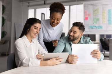 Proje planı üzerinde birlikte çalışan pozitif çok ırklı iş adamları ofis masasında oturup duruyorlar, iletişim için dijital tablet kullanıyorlar, gülümsüyorlar, gülüyorlar.