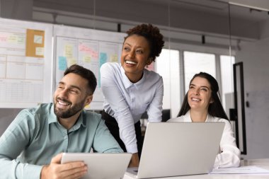 Mutlu çoklu etnik çeşitlilikte genç proje yöneticileri ofis alanında birlikte çalışıyorlar, dizüstü bilgisayar kullanıyorlar, masada buluşuyorlar, beyin fırtınası yapıyorlar, başlarını çeviriyorlar, gülümsüyorlar, gülüyorlar.