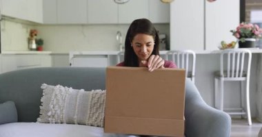Mutlu İspanyol müşteri kadın evde küçük bir karton kutu açıyor, web mağazasından paketleri açıyor, içinde satın alma ile birlikte, gülümsüyor, heyecanlanıyor, şaşırıyor