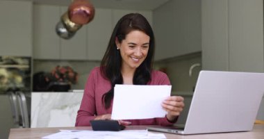 Pozitif serbest muhasebeci kadın proje bütçesini hesaplıyor, kağıt finans raporunu kontrol ediyor, hesap makinesine yazıyor, fatura ödüyor, dizüstü bilgisayarda fatura kesiyor, evden çalışıyor.