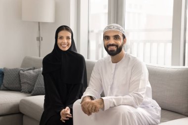 Kameraya poz veren mutlu Arap aile portresi rahat koltukta oturan, siyah abaya giymiş güzel bir kadın, beyaz geleneksel kandura giyen kocası modern evde birlikte vakit geçiriyor.