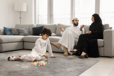 Geleneksel Müslüman kıyafeti giymiş küçük oğlu olan genç bir Arap ailesi boş vakitlerini çağdaş oturma odasında geçiriyor. Çocuk, ailesi koltukta oturmuş konuşurken yerde oynuyor. Oyun zamanı, eğlence zamanı.