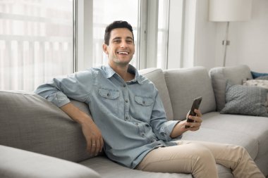 Keyifli genç adam rahat oturma odasında kanepede oturuyor, akıllı telefonunu tutuyor, kameraya gülümsüyor, rahat ve içerik titreşimleri yayıyor, iyi haberlerin ve modern cihaz kullanımının keyfini çıkarıyor.