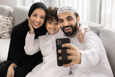 Geleneksel beyaz ve siyah Müslüman kıyafetleri giymiş mutlu Arap çift ve oğlu selfie için poz veriyor, kanepeye oturuyor, akıllı telefon ekranına bakıyor, video görüşmeleri yapıyor ya da fotoğraf çekiyor. Aile anları