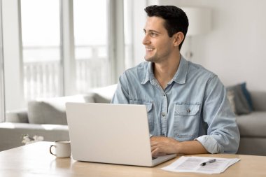 Genç adam dizüstü bilgisayarla masada oturuyor, gözünü kaçırıyor, uzak bir işte çalışıyor ya da yaratıcı görevlerle uğraşıyor, tamamlıyor, gülümsüyor, işten tatmin oluyor, yeni mesleki başarılar veya fırsatlar düşünüyor.