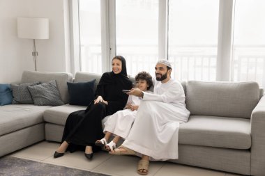 Genç Ortadoğulu aile, geleneksel Müslüman kıyafetleriyle modern oturma odasındaki kanepede oturup film ya da çizgi film seçen, favori komedi dizilerinin ya da dizilerin keyfini çıkarıyorlar.