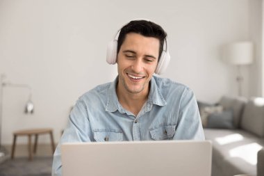 Gülümseyen İspanyol adam internet içeriğini izleyen kablosuz kulaklık takıyor, kişisel ya da iş dünyası tarafından yapılan video konferans toplantısına katılıyor, gülümsüyor, haberleri paylaşıyor, evde video uygulaması ve dizüstü bilgisayar kullanıyor.