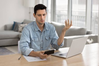 Genç işadamı, kafası karışmış, hayal kırıklığına uğramış ifade ile hesap makinesine bakıyor, mali konularda sorunlar yaşıyor, zorlu bütçelerle uğraşıyor, beklenmedik harcamalar, finansal planlardaki tutarsızlıklar