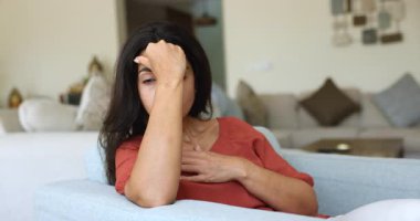 Depresyondaki genç Latin kadın kendini kötü hissediyor, stresli, üzgün, başıyla göğsüne dokunuyor, koltuğunda oturuyor, baş ağrısı, depresyon, ilgisizlik, kaybı düşünüyor, başarısızlığı düşünüyor.