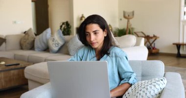 Evde serbest çalışan genç Latin bir kadın, dizüstü bilgisayar kullanıyor, uzak iş görevleri için iletişim kuruyor, rahat koltukta dinleniyor, bilgisayarını kucağında tutuyor.