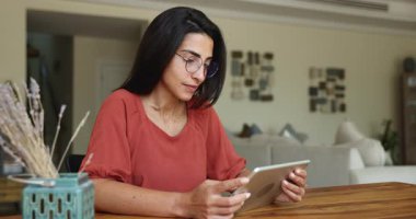 İletişim masasında tablet bilgisayar kullanarak, online medya içeriğini izleyerek, mutlu olarak, gülerek, ev içi internet iletişiminden zevk alarak, şık gözlüklü, genç İspanyol bir kadın.